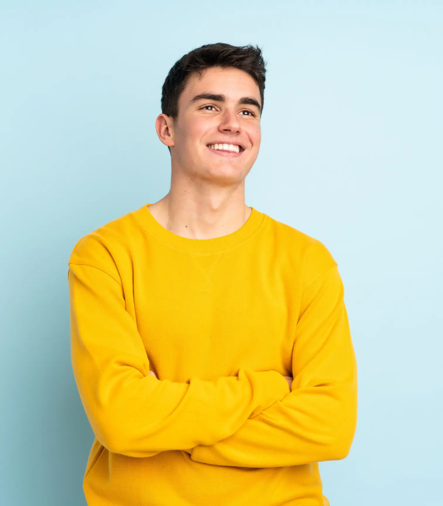 An older teenage boy with a yellow sweater against a blue backdrop crossing his arms and smiling