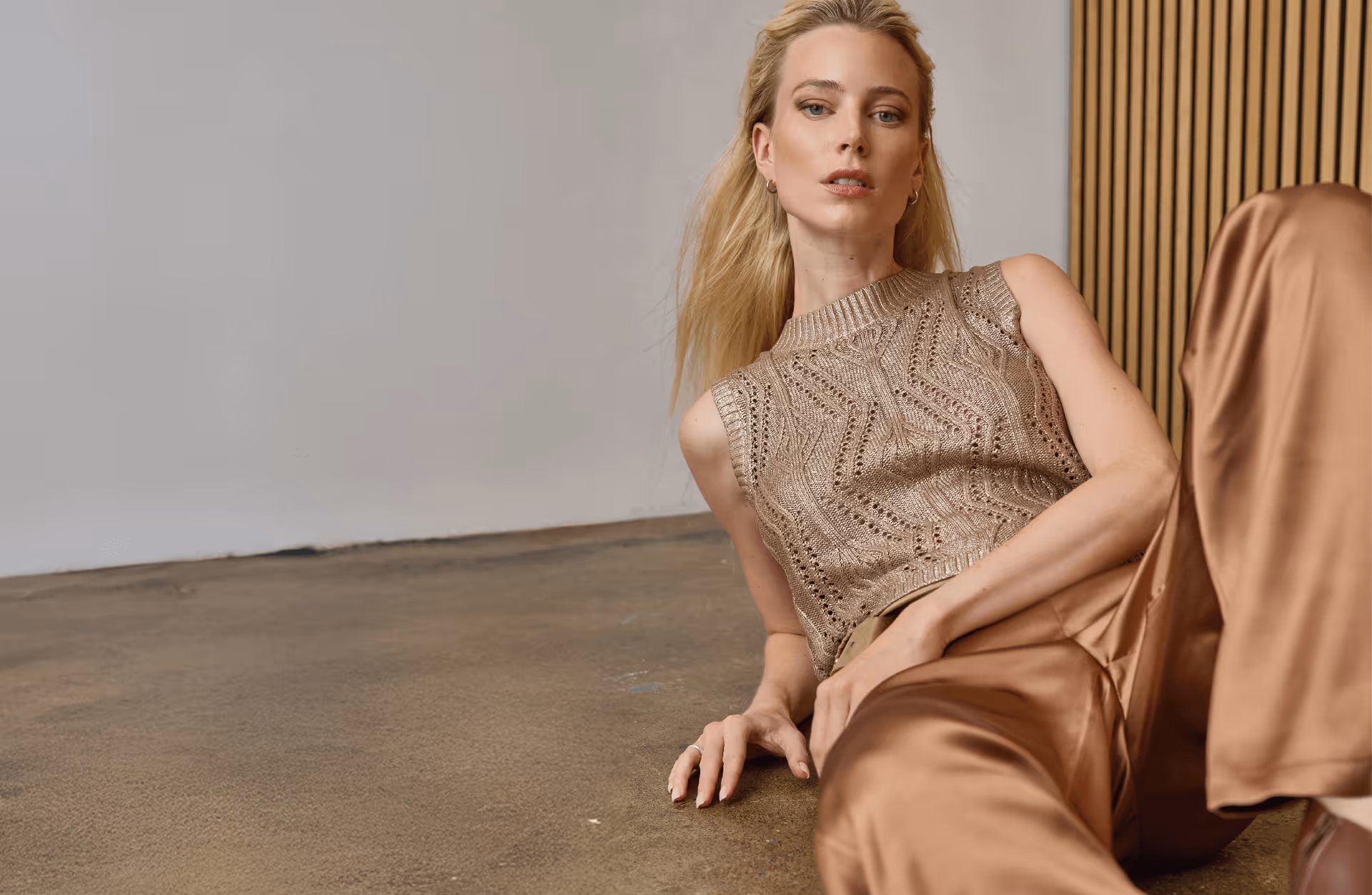 Blonde woman reclining on a concrete floor wearing a sleeveless knitted beige top and shiny brown satin pants.