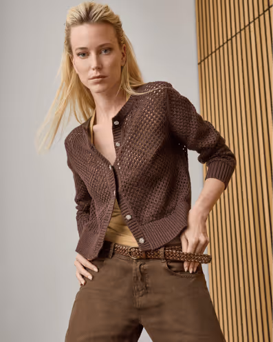 Blonde woman wearing a brown knit cardigan over a beige top with brown pants and a braided belt posing indoors.