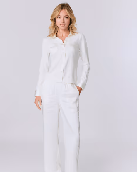 Blonde woman wearing a white button-up long-sleeve shirt and white pants standing against a light gray background.