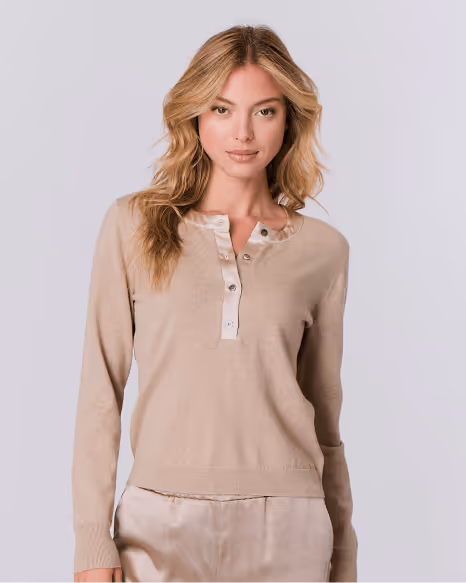 Woman with long blonde hair wearing a beige long-sleeve sweater with button details and light beige pants.