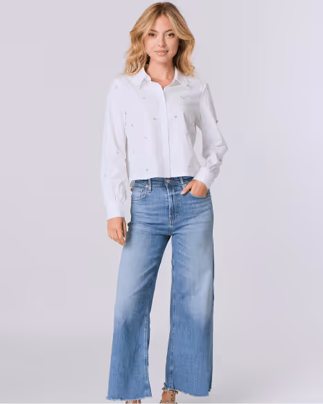Woman with blonde hair wearing a white button-up blouse and wide-leg blue jeans standing against a light gray background.