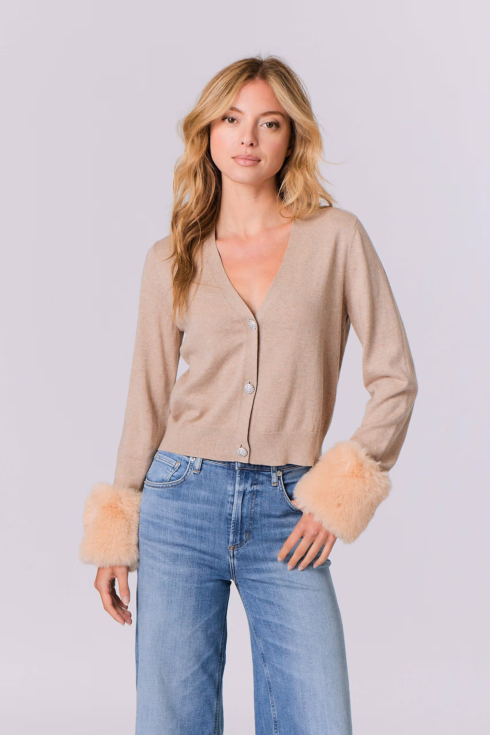 Blonde woman wearing a beige cardigan with furry cuffs and light blue high-waisted jeans.