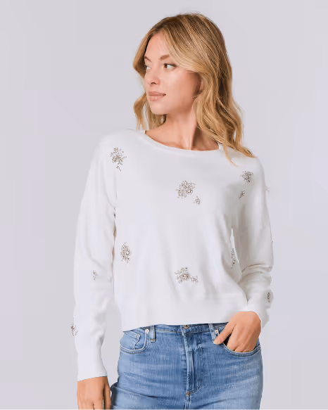 Woman wearing a white sweater with small bead embellishments paired with blue jeans, looking to the side.
