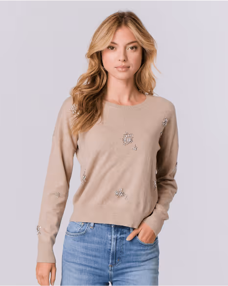 Blonde woman wearing a beige sweater with decorative gems and blue jeans, posing with one hand in her pocket.