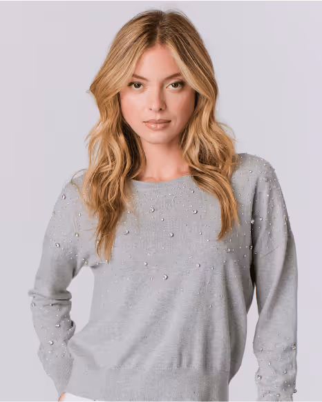 Blonde woman wearing a light gray sweater adorned with small pearls, posing against a plain light background.