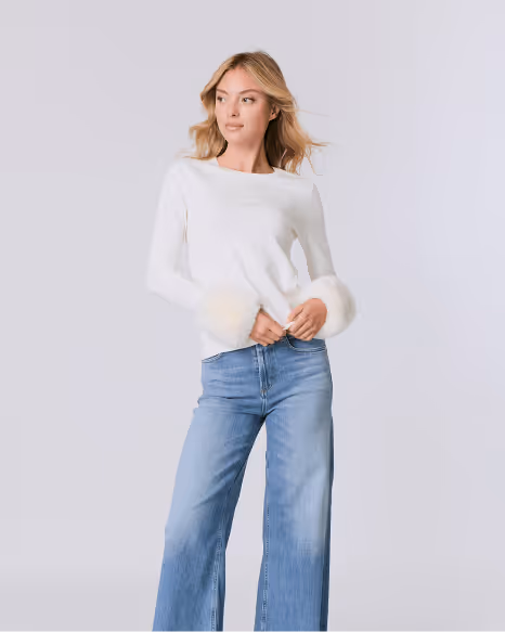 Woman wearing a white long-sleeve top with fluffy cuffs and blue wide-leg jeans, standing against a plain background.