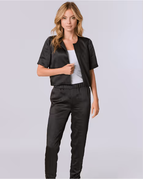 Blonde woman wearing a black short-sleeve jacket over a white top and matching black pants against a light gray background.