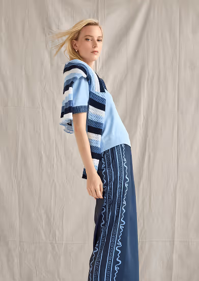 Blonde woman wearing a light blue shirt with a blue striped sweater draped over her shoulders and navy blue pants with light blue patterns.