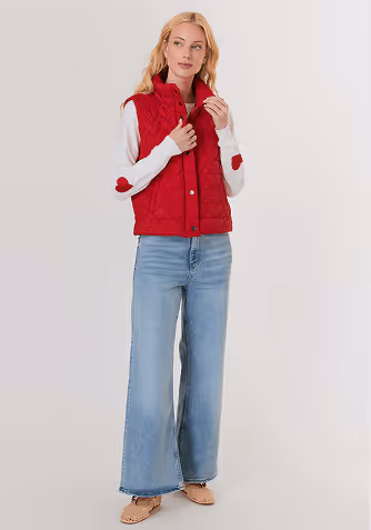 Woman with blonde hair wearing a white button-up blouse and wide-leg blue jeans standing against a light gray background.