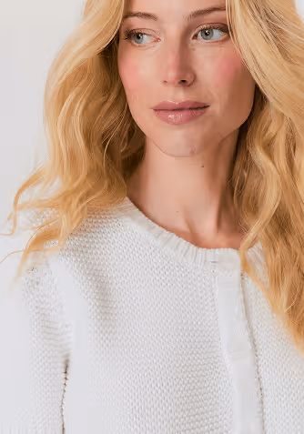 Blonde woman wearing a beige sweater with decorative gems and blue jeans, posing with one hand in her pocket.