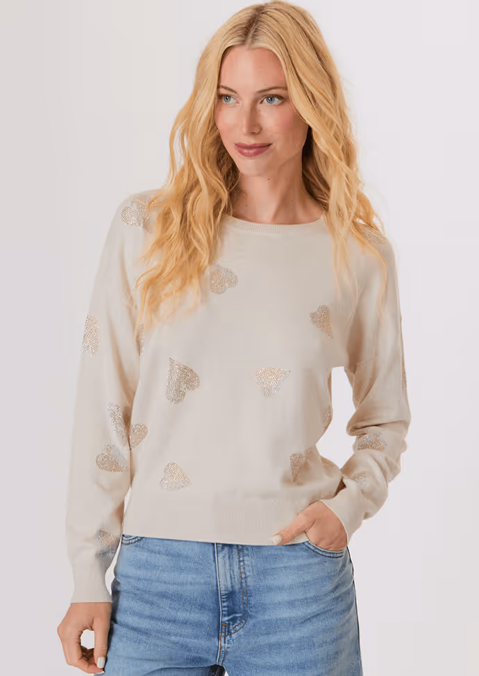 Blonde woman wearing a white button-up shirt with pockets and white pants, posing with hands in pockets against a light background.