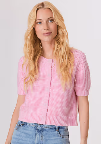 Woman wearing a white long-sleeve top with fluffy cuffs and blue wide-leg jeans, standing against a plain background.