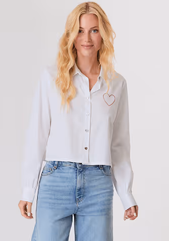 Blonde woman in a navy button-up shirt with embellished collar and light blue jeans against a plain background.