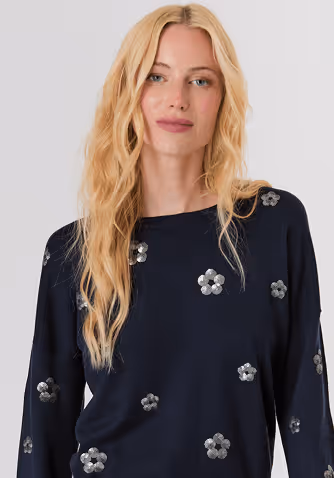 Blonde woman wearing a red knit sweater and white pants standing against a plain light background.