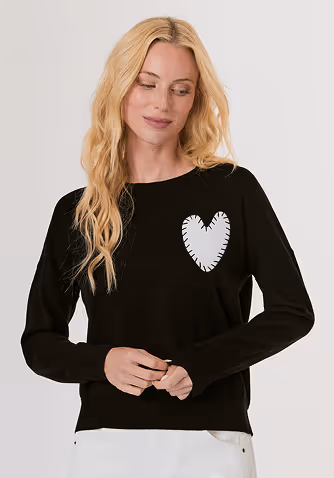 Blonde woman wearing a black short-sleeve open jacket over a white sleeveless top and white pants, standing against a light gray background.