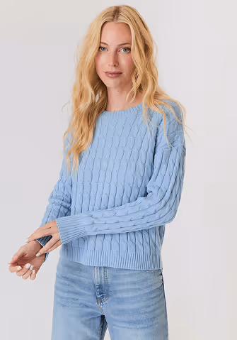 Blonde woman wearing a black and white sweater with a snowflake pattern and blue jeans standing against a light gray background.