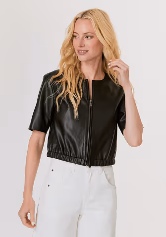 Blonde woman wearing a black short-sleeve jacket over a white top and matching black pants against a light gray background.