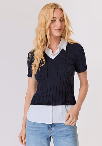 Blonde woman in a navy blue sweater and blouse touching her hair against a light gray background.