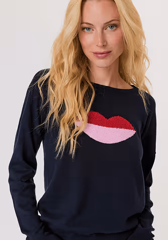 Blonde woman wearing a beige long-sleeve sweater with a pleated hem and blue jeans against a plain light gray background.