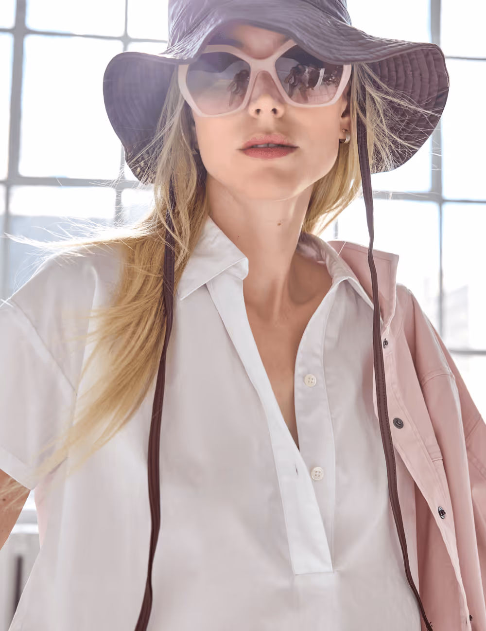 Blonde woman wearing a wide-brimmed hat, large sunglasses, a white shirt, and a pink jacket draped over one shoulder.