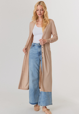 Woman with long blonde hair wearing a sleeveless beige knitted top and shiny light brown wide-leg pants, standing with arms crossed.