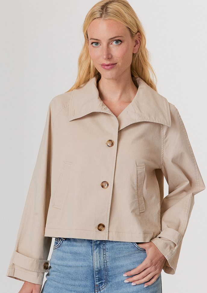 Woman with blonde hair wearing a white buttoned cardigan with decorative buttons and light blue jeans against a plain light background.