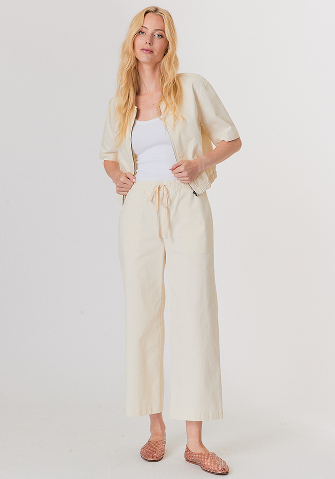 Blonde woman wearing a white button-up long-sleeve shirt and white pants standing against a light gray background.