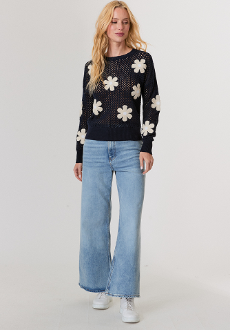 Blonde woman in a navy blue sweater and blouse touching her hair against a light gray background.