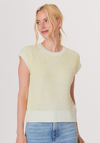 Blonde woman wearing a black and white sweater with a snowflake pattern and blue jeans standing against a light gray background.