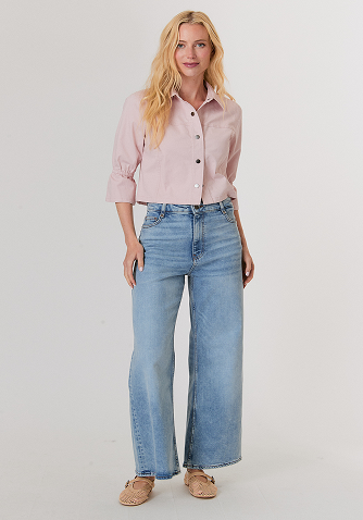 Blonde woman wearing a red knit sweater and white pants standing against a plain light background.