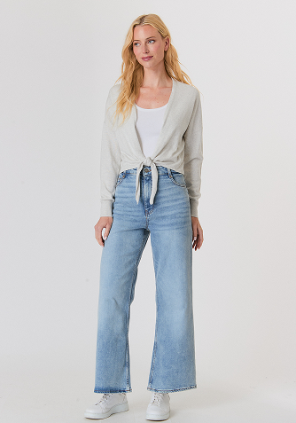 Woman wearing a white long-sleeve top with fluffy cuffs and blue wide-leg jeans, standing against a plain background.