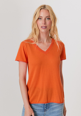 Blonde woman wearing a red knit sweater and white pants standing against a plain light background.