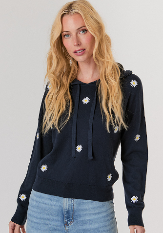 Blonde woman wearing a black short-sleeve open jacket over a white sleeveless top and white pants, standing against a light gray background.