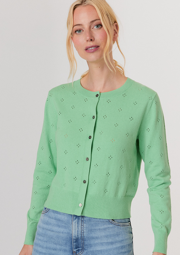 Blonde woman wearing a white button-up shirt with pockets and white pants, posing with hands in pockets against a light background.