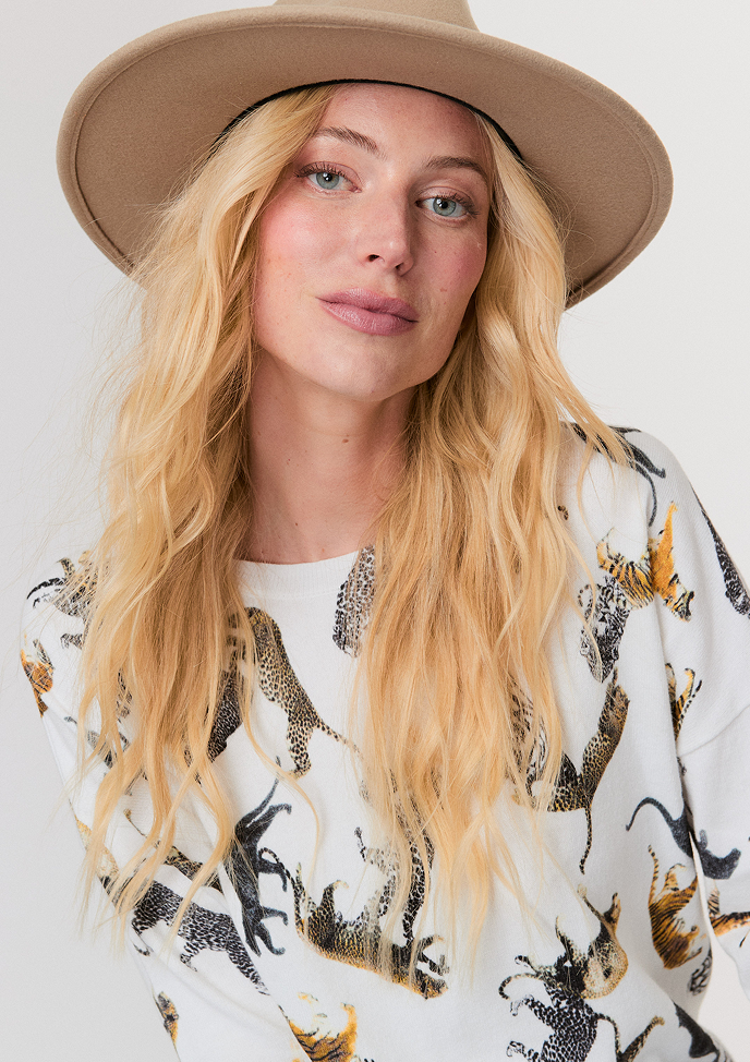 Blonde woman wearing a white button-up shirt with pockets and white pants, posing with hands in pockets against a light background.