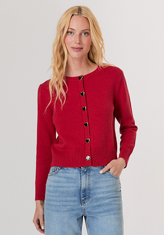 Blonde woman wearing a red knit sweater and white pants standing against a plain light background.