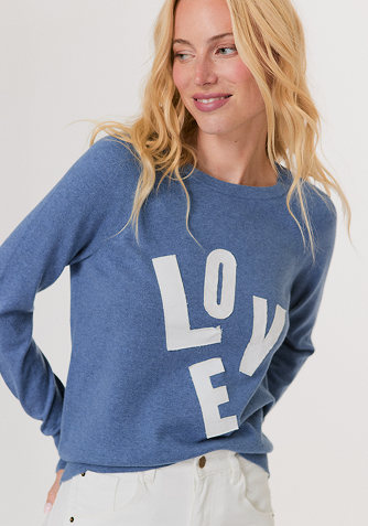 Woman wearing a white long-sleeve top with fluffy cuffs and blue wide-leg jeans, standing against a plain background.