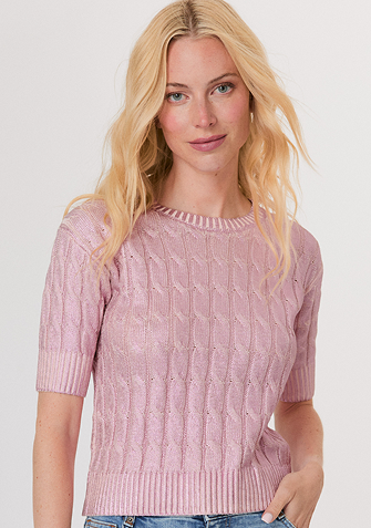 Blonde woman wearing a black and white sweater with a snowflake pattern and blue jeans standing against a light gray background.