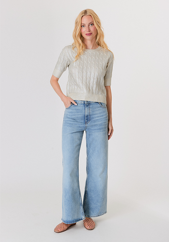 Blonde woman wearing a black short-sleeve open jacket over a white sleeveless top and white pants, standing against a light gray background.