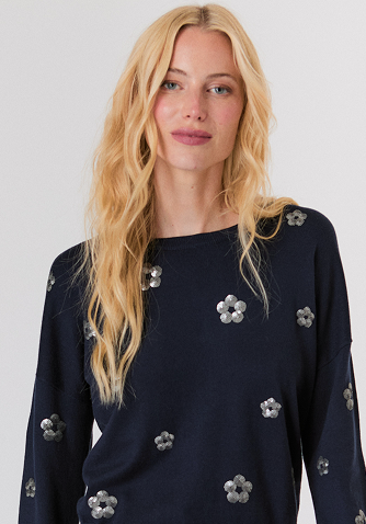 Blonde woman in a navy button-up shirt with embellished collar and light blue jeans against a plain background.