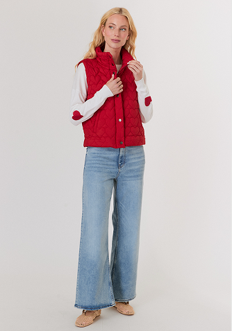 Blonde woman wearing a black short-sleeve open jacket over a white sleeveless top and white pants, standing against a light gray background.