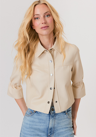 Blonde woman wearing a black short-sleeve open jacket over a white sleeveless top and white pants, standing against a light gray background.