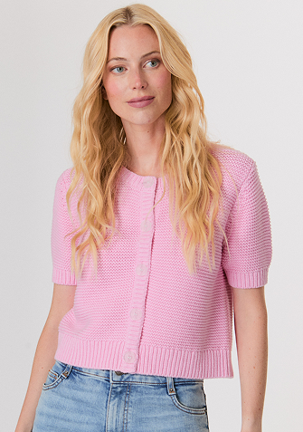 Blonde woman wearing a black and white sweater with a snowflake pattern and blue jeans standing against a light gray background.