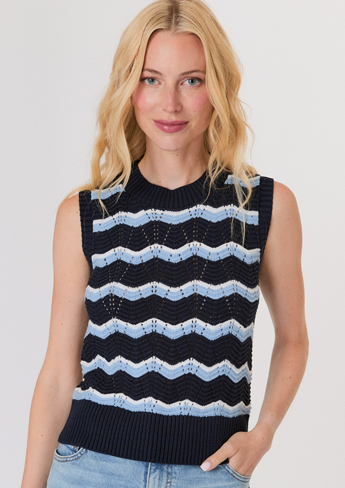 Blonde woman wearing a white button-up shirt with pockets and white pants, posing with hands in pockets against a light background.