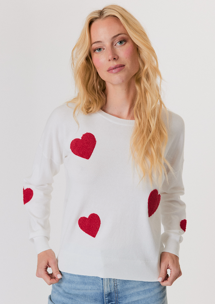 Blonde woman wearing a white button-up shirt with pockets and white pants, posing with hands in pockets against a light background.