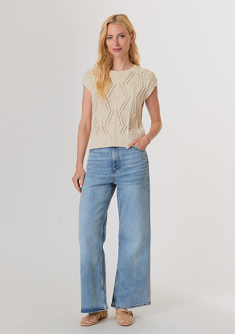 Blonde woman wearing a black and white sweater with a snowflake pattern and blue jeans standing against a light gray background.