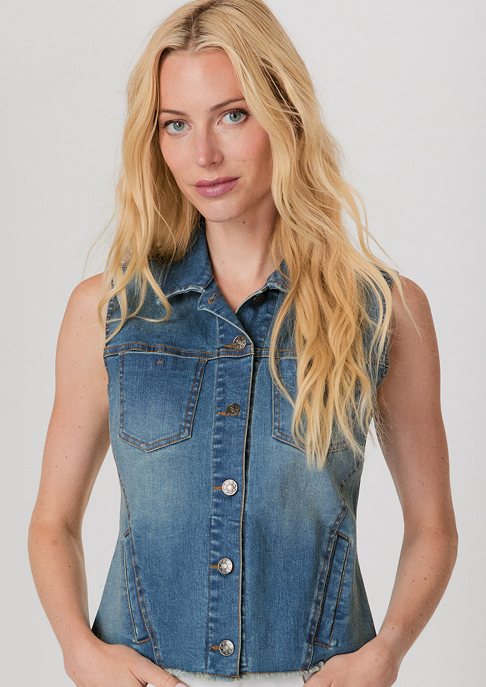 Blonde woman wearing a white button-up shirt with pockets and white pants, posing with hands in pockets against a light background.