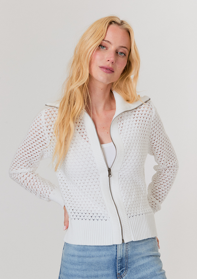 Blonde woman wearing a white button-up shirt with pockets and white pants, posing with hands in pockets against a light background.