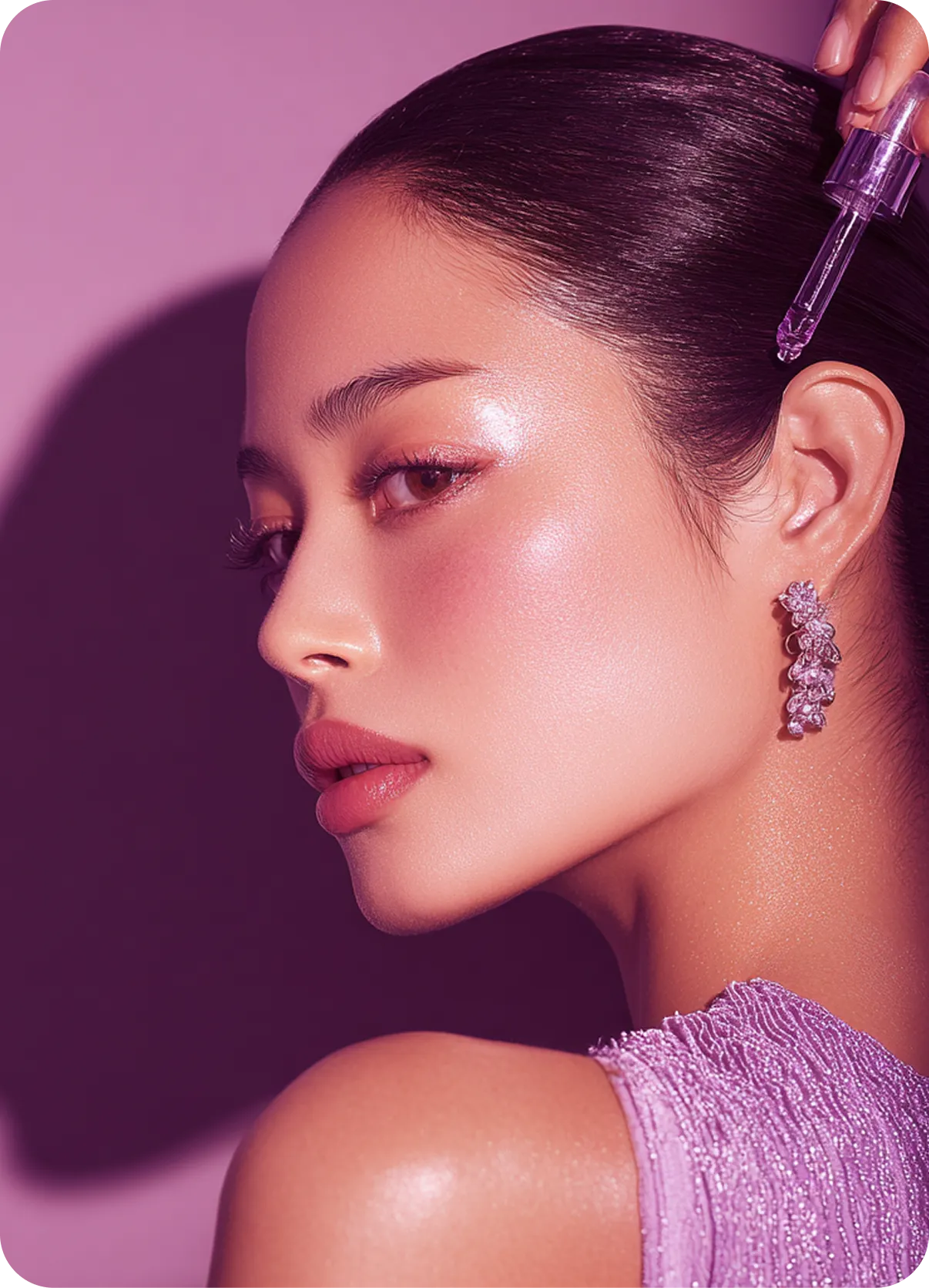 Close-up of a woman with glowing skin and purple shimmer applying a dropper serum near her temple, wearing purple earrings and a textured lavender top.